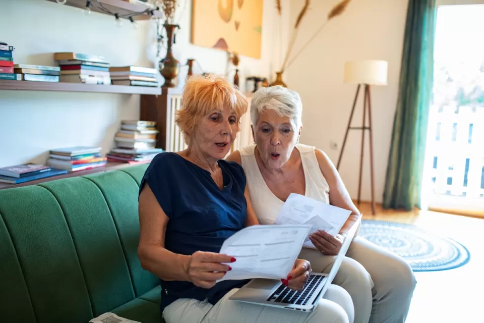 Two older women sitting on green sofa looking at paperwork or bills.